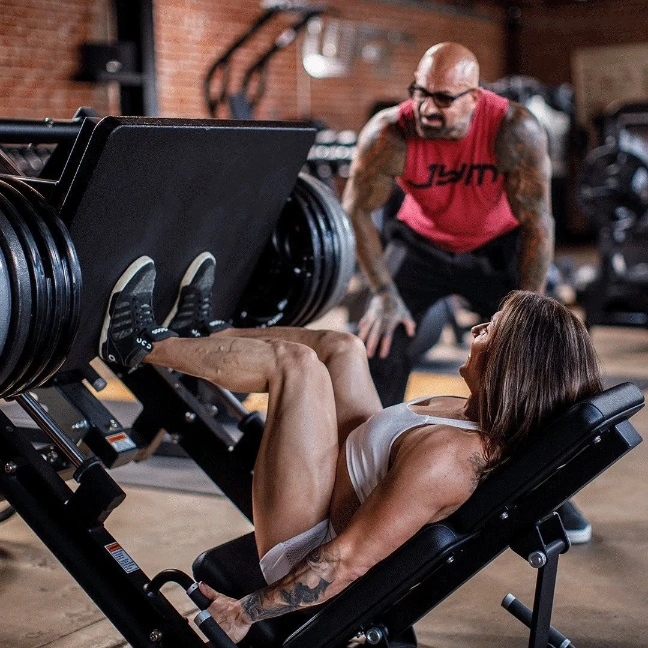 A woman in workout attire performs a leg press at the gym, pushing a heavy weight stack with her legs. A muscular personal trainer in a red sleeveless top stands nearby, providing encouragement and guidance to help her achieve her fitness goals. The gym equipment and brick walls are visible in the background.