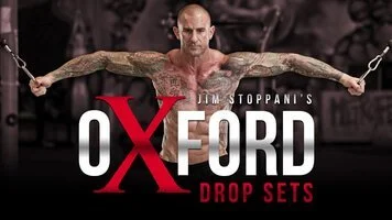 A muscular, tattooed man performs a cable crossover exercise in a gym. Large text in front of him reads, "Oxford Drop Sets," with "Jim Stoppani's" above it. The background is blurred gym equipment, emphasizing personalized training programs tailored to fitness goals.